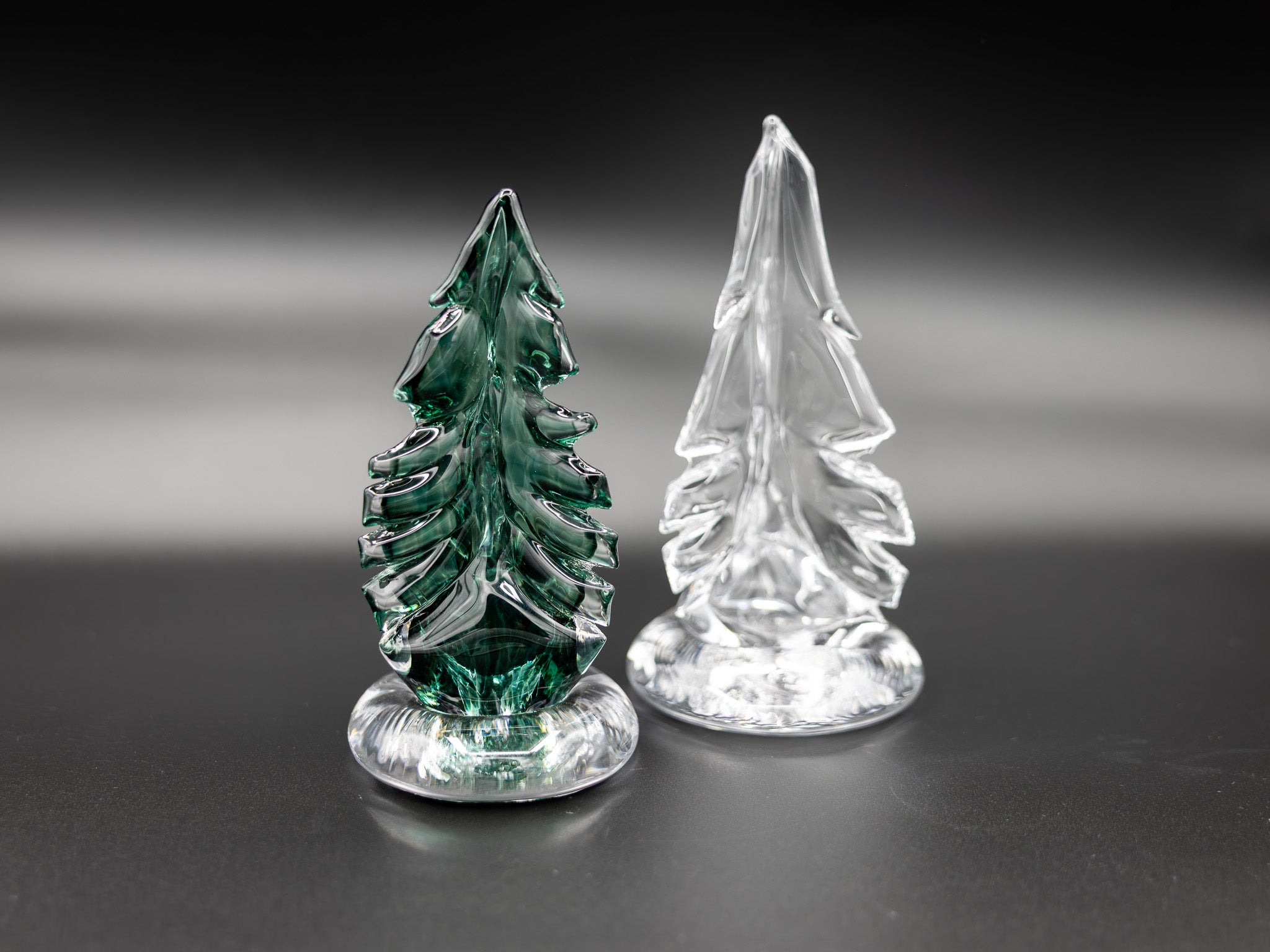Two small glass trees, one green and one clear, on a reflective surface with a dark background.
