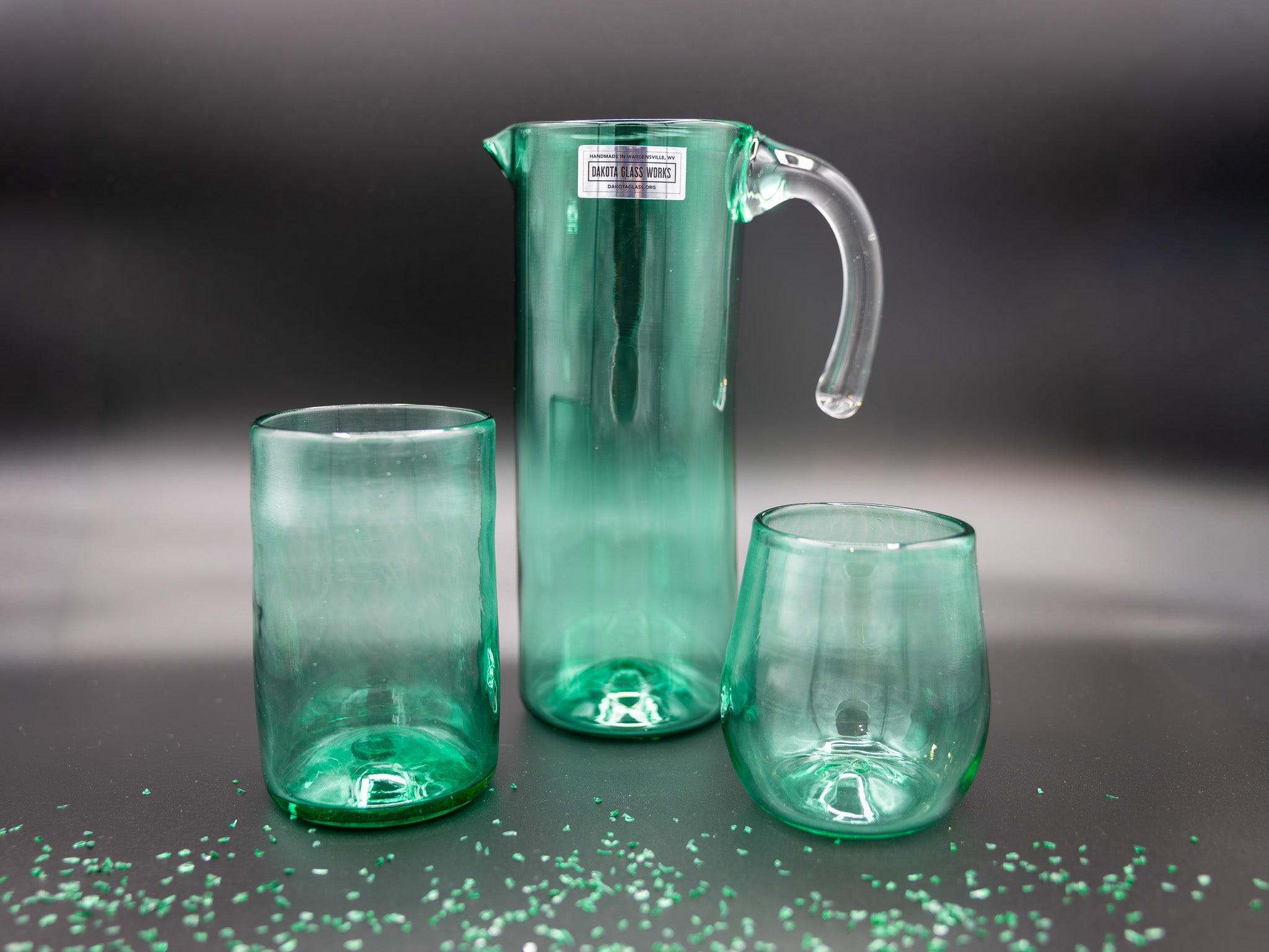 Green glass pitcher and two glasses on a dark surface with a blurred background