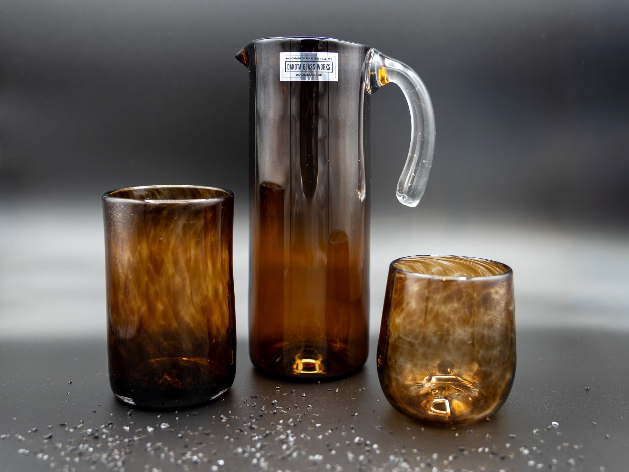 A brown stemless wine glass, brown tumbler and a matching pitcher on a dark surface with a blurred background.