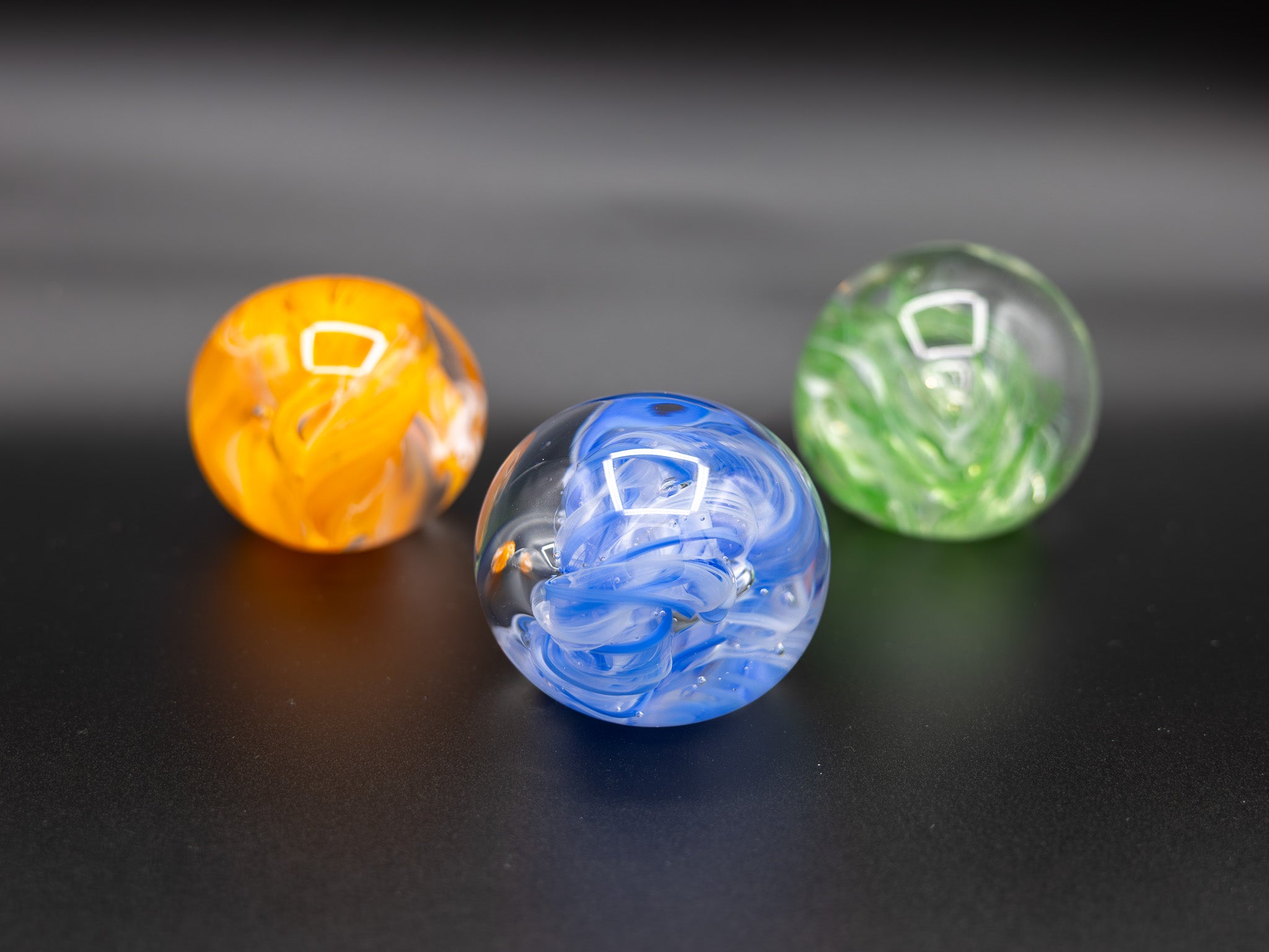 Three glass paperweights in orange, blue, and green on a dark background