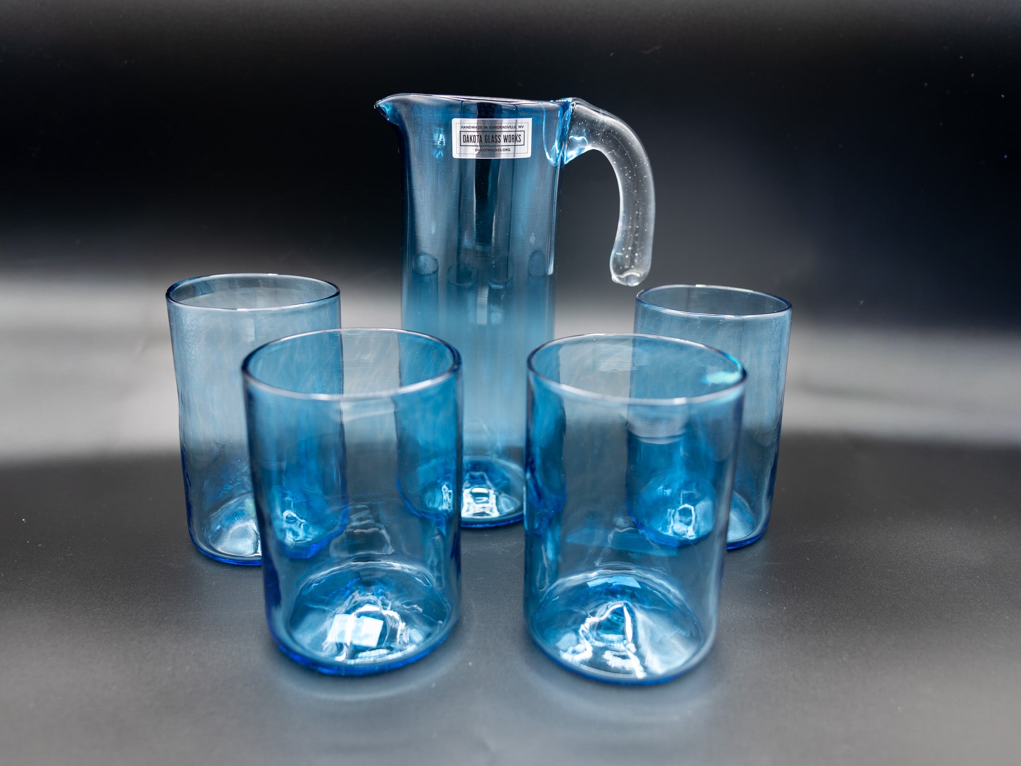 Set of blue glass tumblers and a pitcher on a reflective surface with a dark background