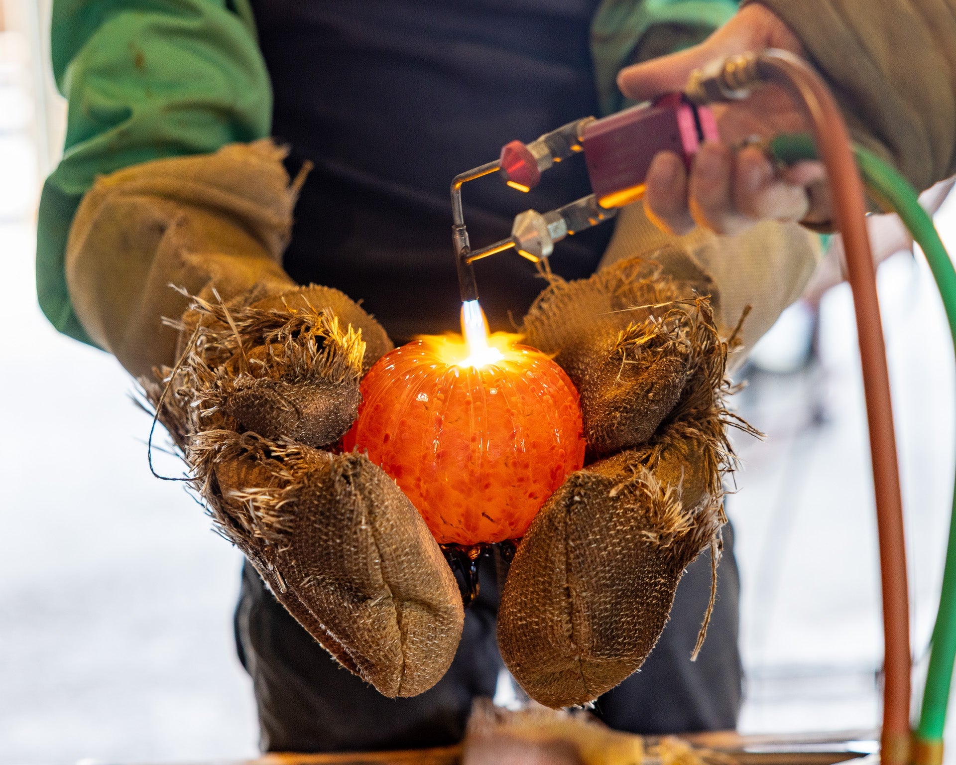A person wearing thick brown burlap work gloves holds a small orange pumpkin while using a torch lighter with a visible flame. The person also wears a green long-sleeve shirt and a dark apron, with a hose connected to the lighter in the background.