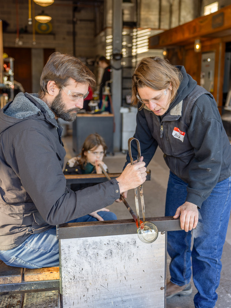 a father and daughter working with a glassblower to create a cup 