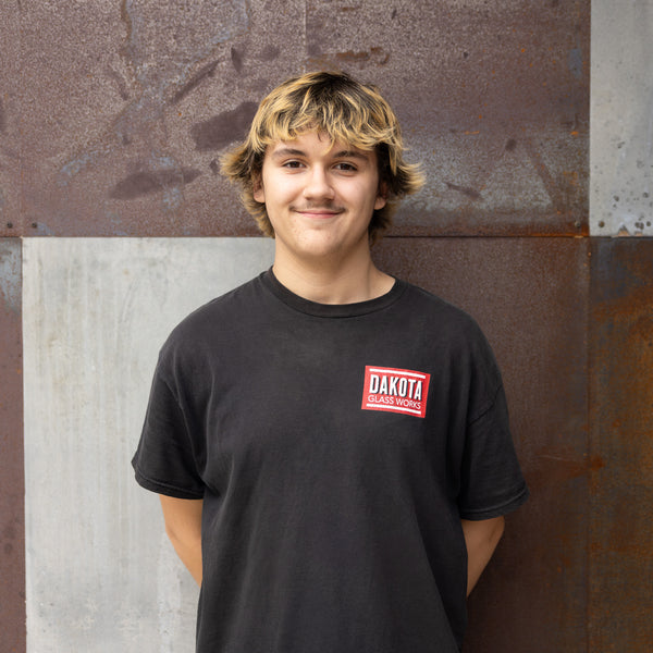 Person wearing a black t-shirt with 'Dakota Glass Works' logo against a textured wall.