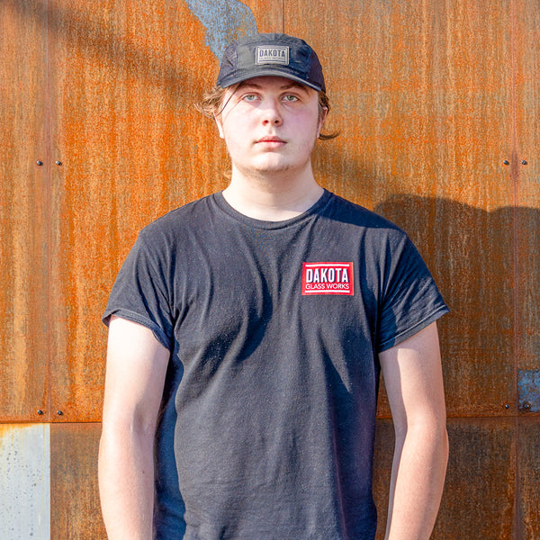 Person wearing a dark t-shirt with a Dakota Glass Works logo against a rusty metal background