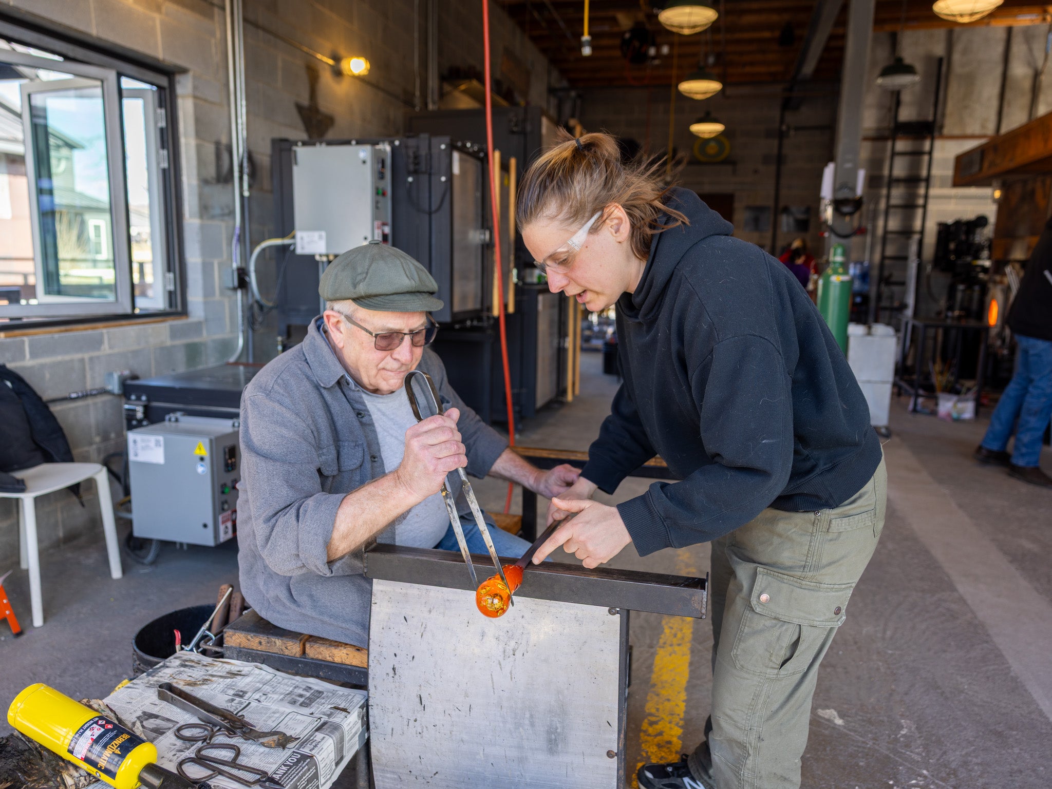 Two people working with glassblowing equipment in a workshop.