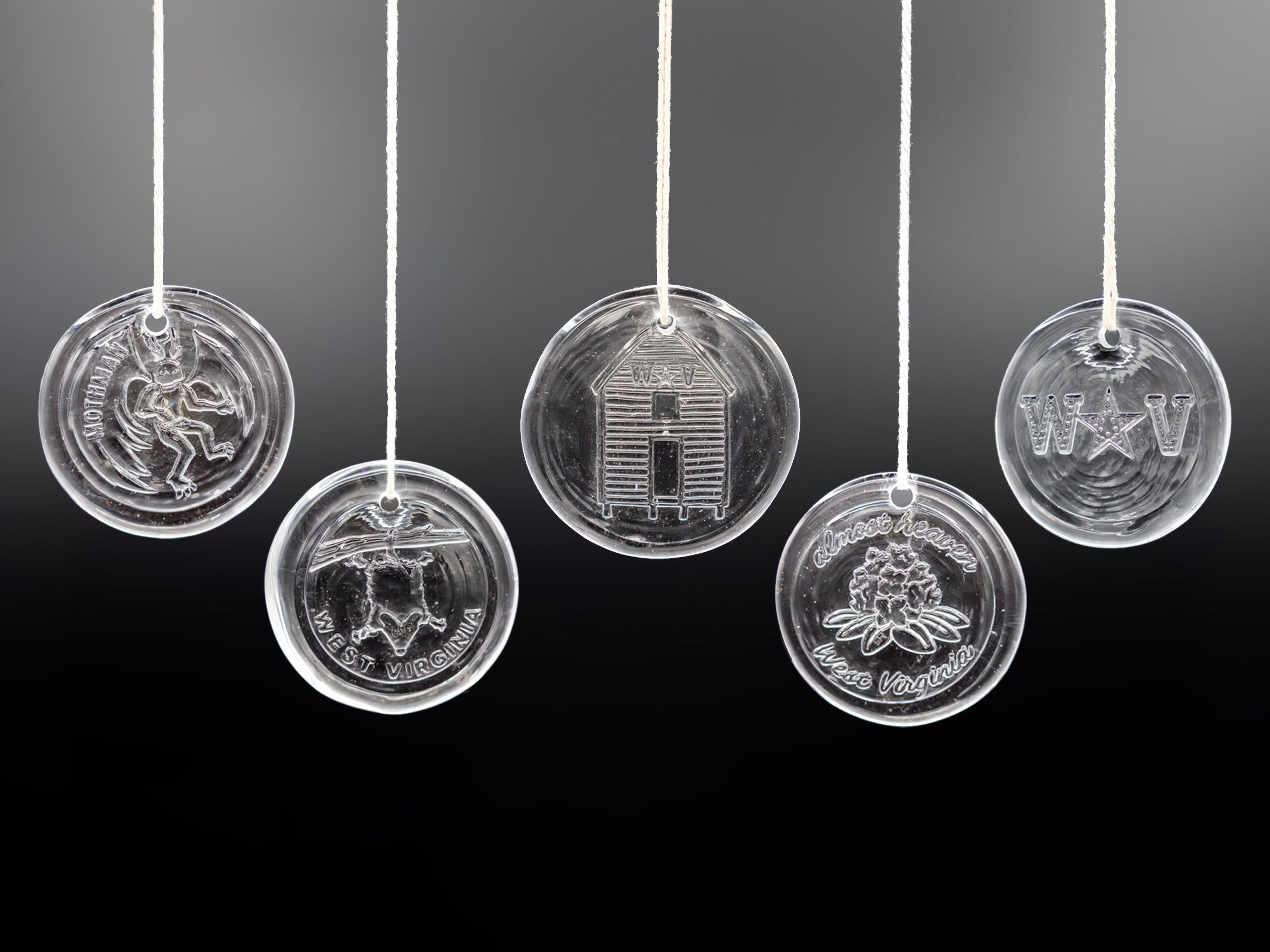 Five clear glass hanging ornaments with engraved designs, suspended by white strings against a dark gradient background.