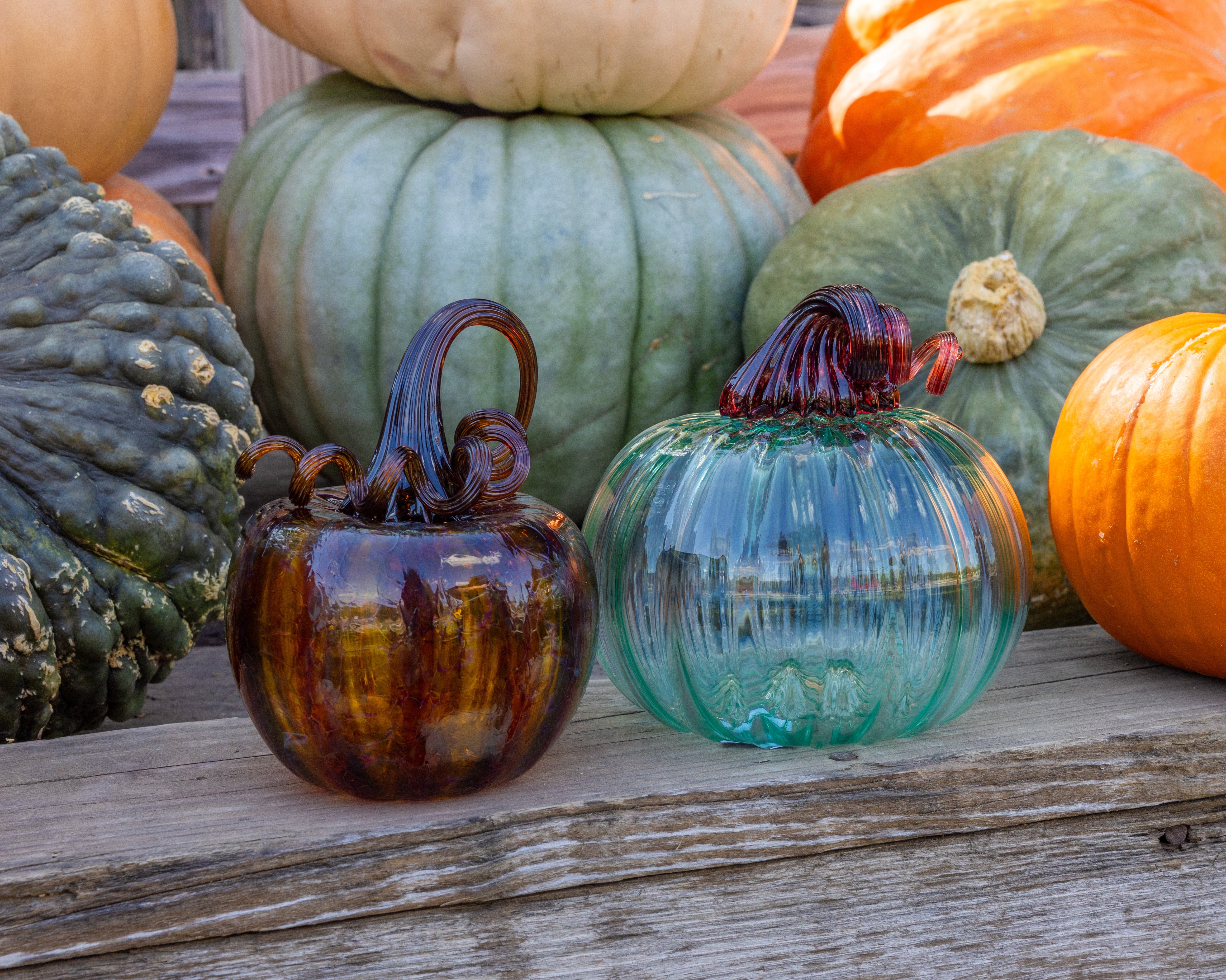 Two decorative glass pumpkins, one amber-brown with a dark red top and one translucent aqua-blue with a dark red top, displayed on a weathered wooden surface surrounded by real pumpkins.