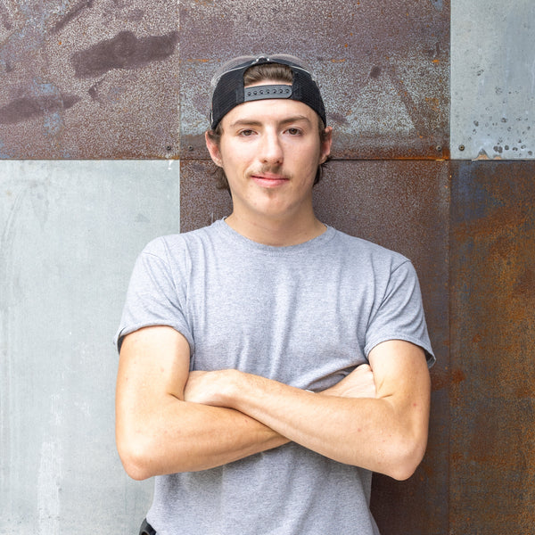 Person wearing a gray t-shirt and black cap standing against a textured wall.