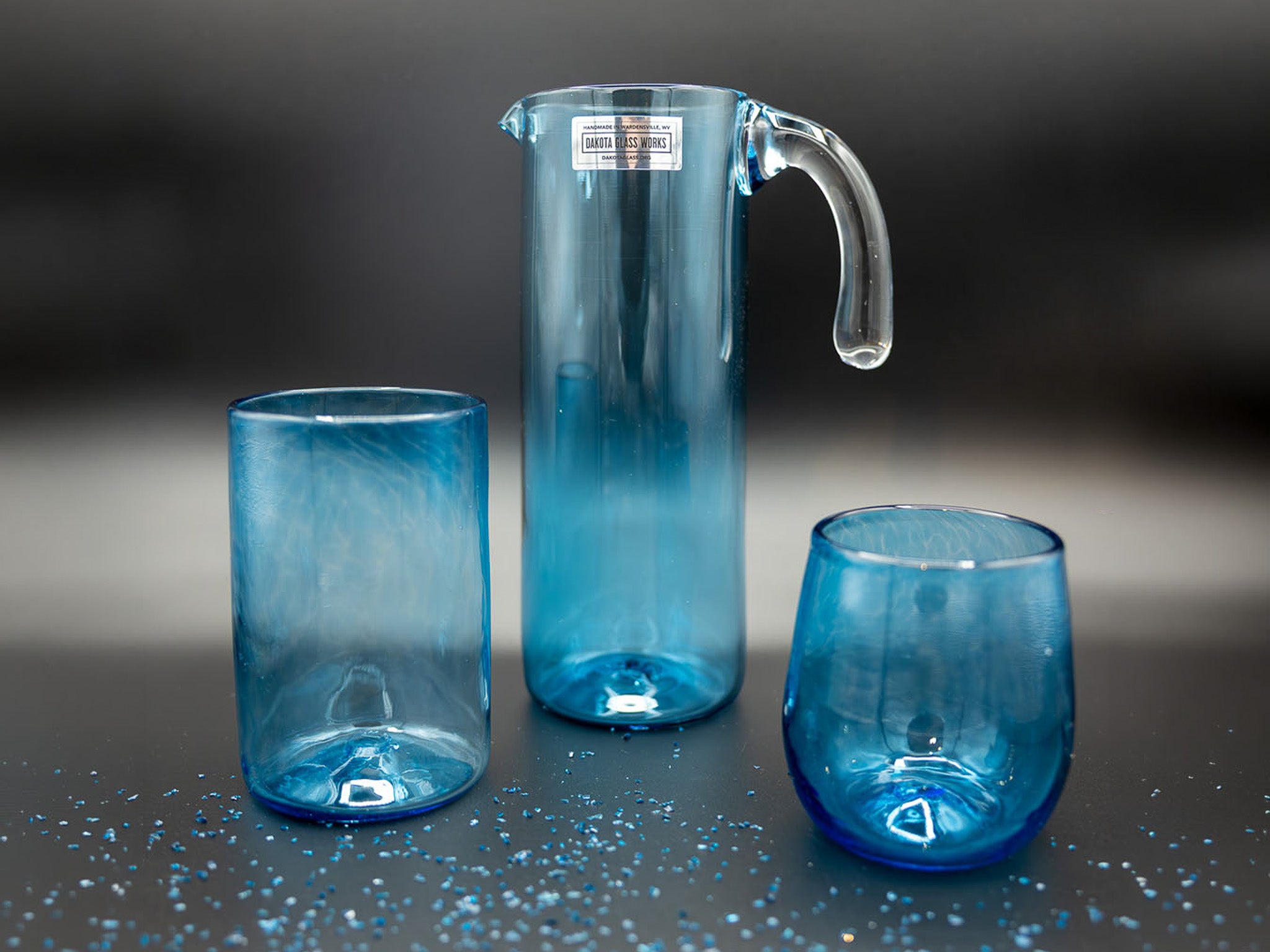 Set of blue glassware including a pitcher. a tumbler, and a stemless wine glass on a dark surface.